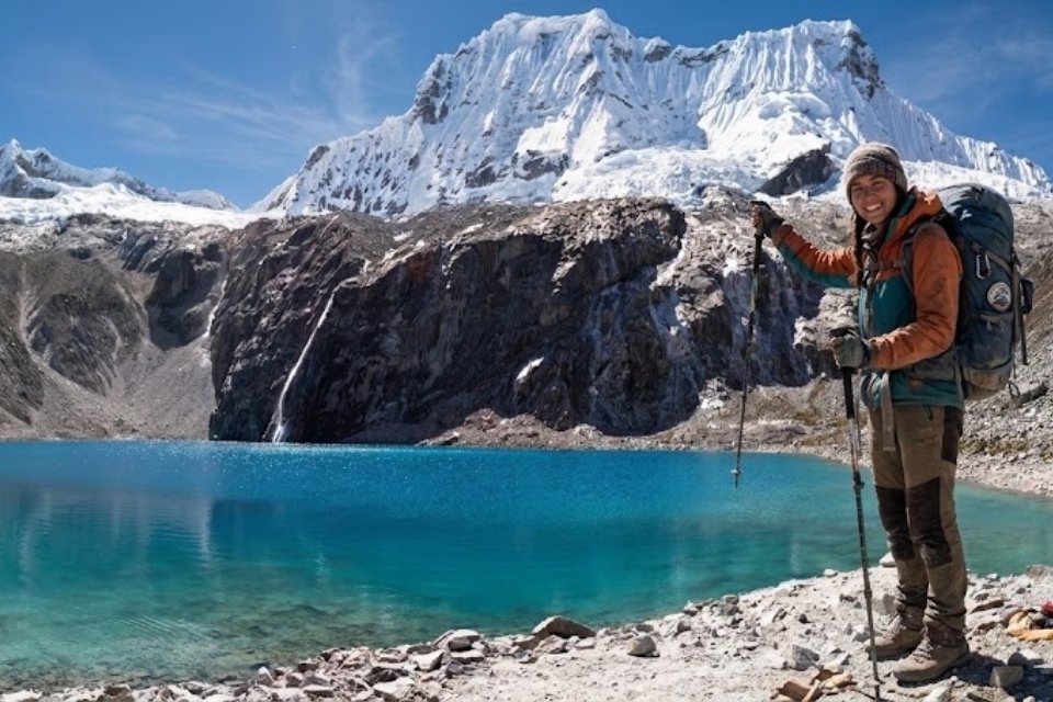 A happy hiker smiling at the shore of Laguna 69 in Huaraz, fully equipped with trekking poles and a backpack, standing before the vivid turquoise water and the massive snow-capped Chacraraju peak on a professional guided tour with Andeando Peru.