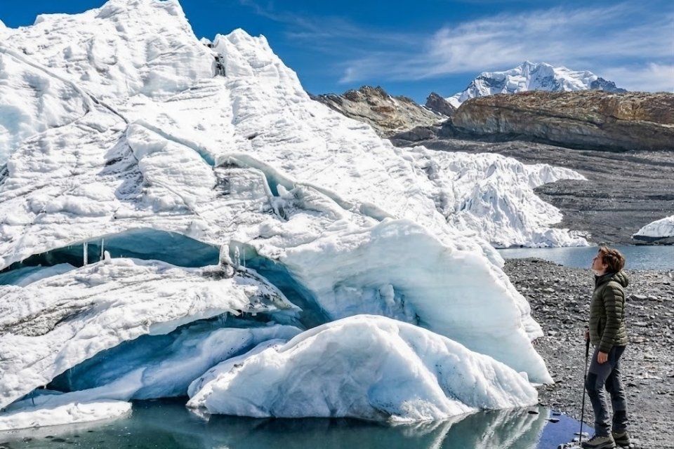 A female hiker standing at the edge of the blue-tinted Pastoruri Glacier ice wall in the Cordillera Blanca. The scene shows the vast scale of the melting glacier at 17,224 feet, featuring a glacial lagoon and rugged Andean peaks under a clear sky. Photo by Andeando Peru.