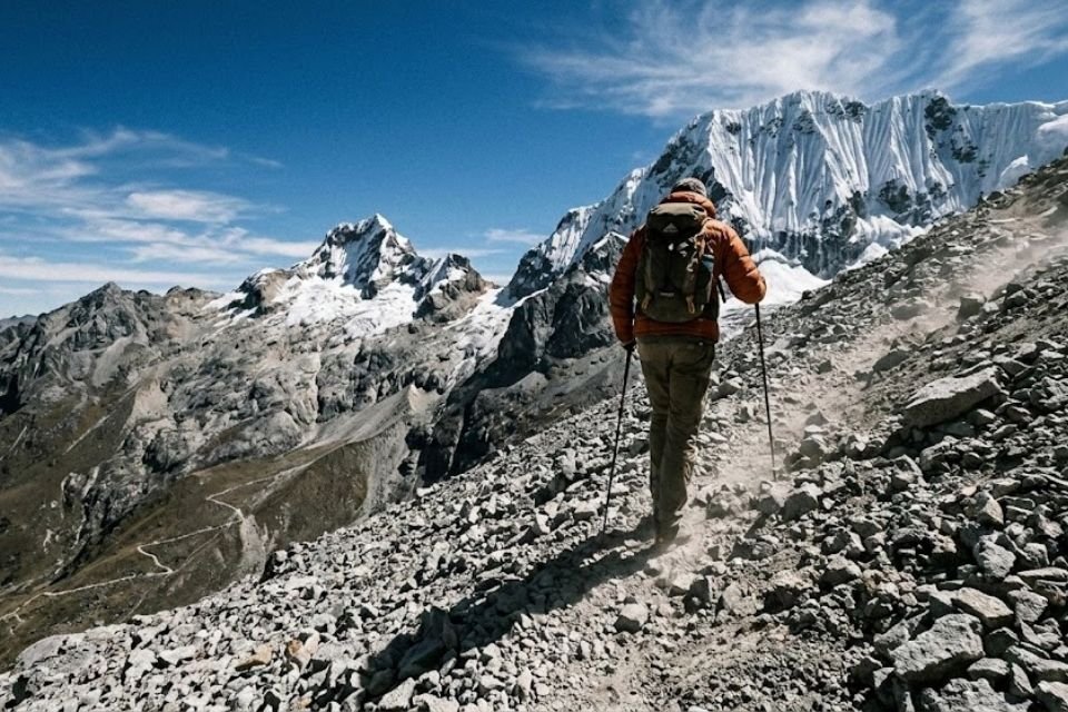 A hiker using trekking poles to navigate a steep, dusty scree slope in the Cordillera Blanca near Huaraz, with massive glaciated Andean peaks in the background under a clear blue sky, demonstrating professional mountain training by Andeando Perú.